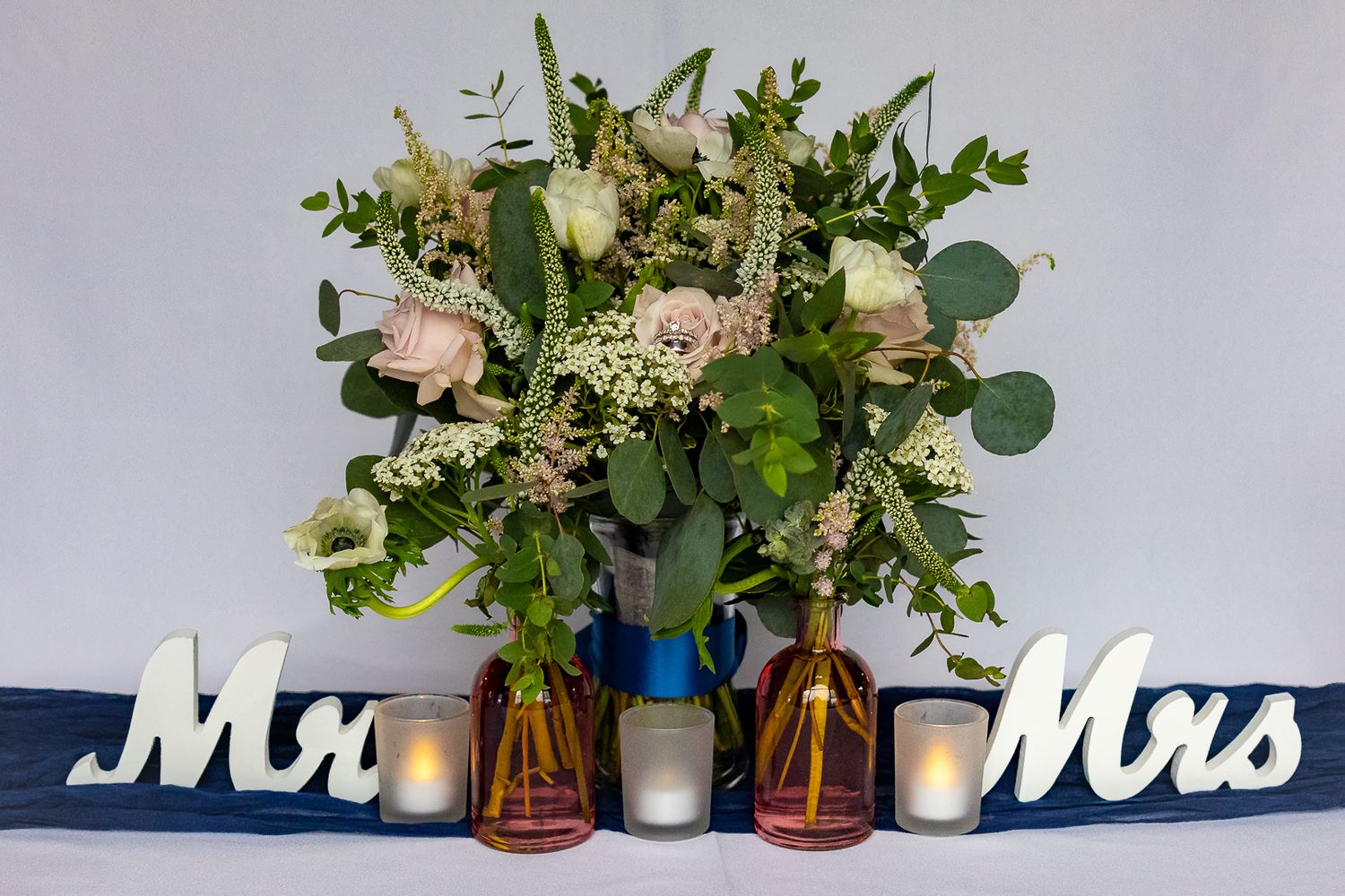 Wedding table decor with Mr and Mrs signs, pink roses bouquet, eucalyptus greenery, and glowing candles in amber vases.