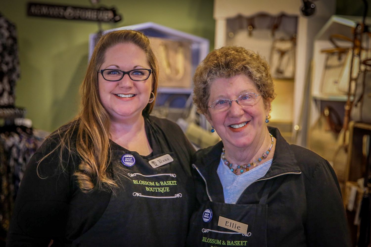 Two smiling women in matching black Blossom & Basket Boutique uniforms pose together inside a charming retail shop.