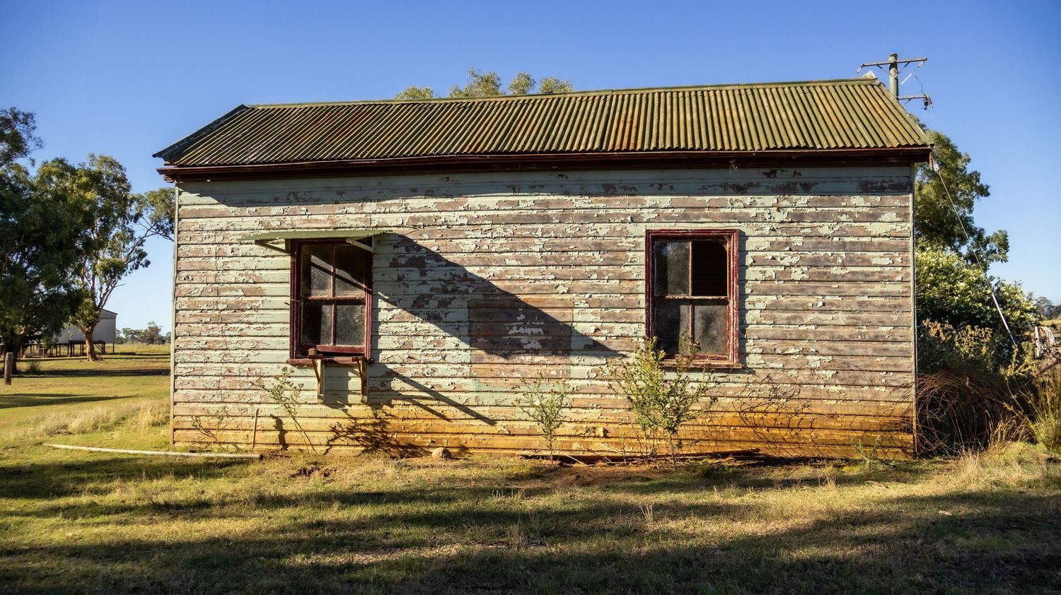 An abandoned weathered wooden house with a corrugated iron roof sits on a dry grassy field under a blue sky.