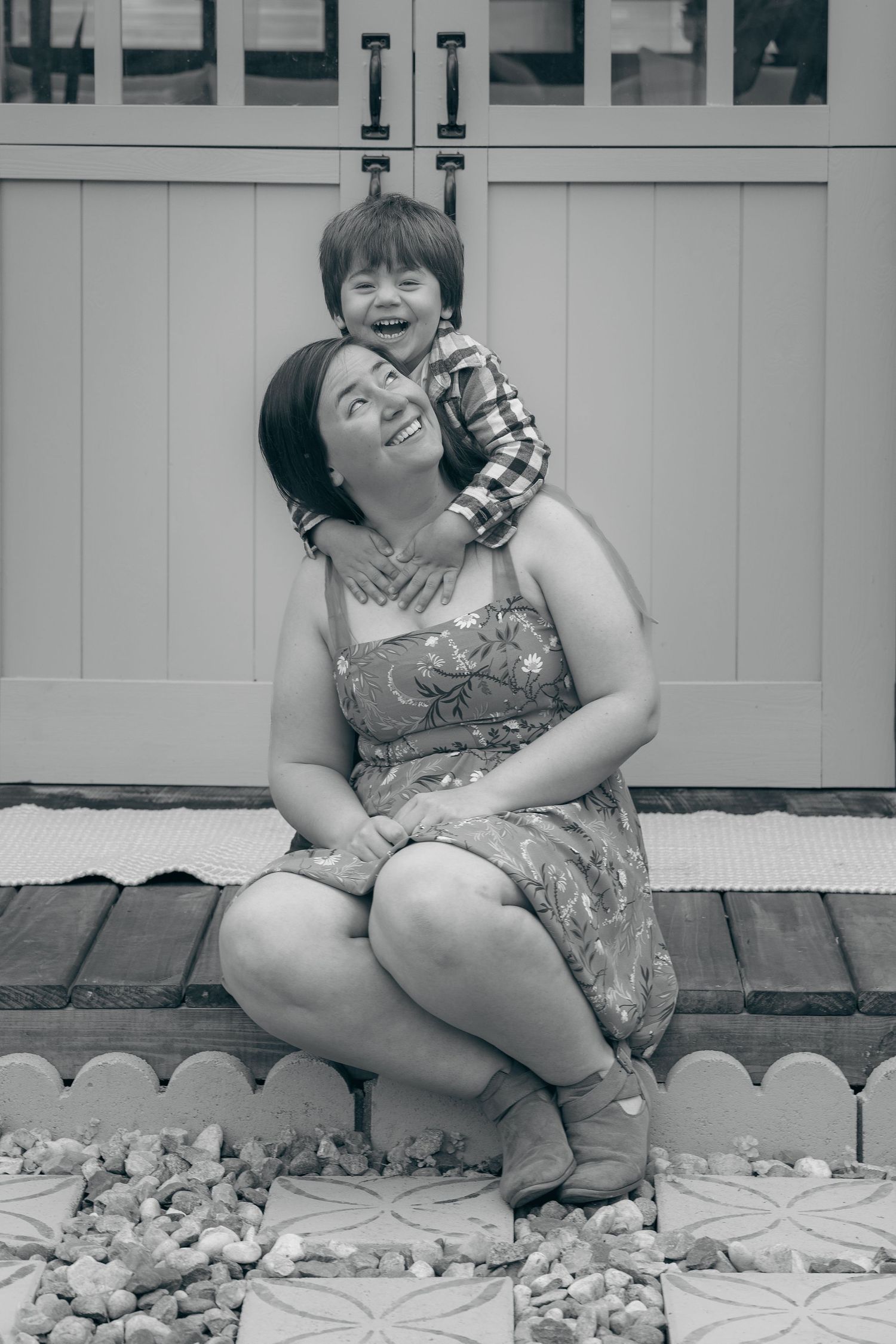 Black and white photo of a smiling mother sitting on steps with her young son hugging her from behind.