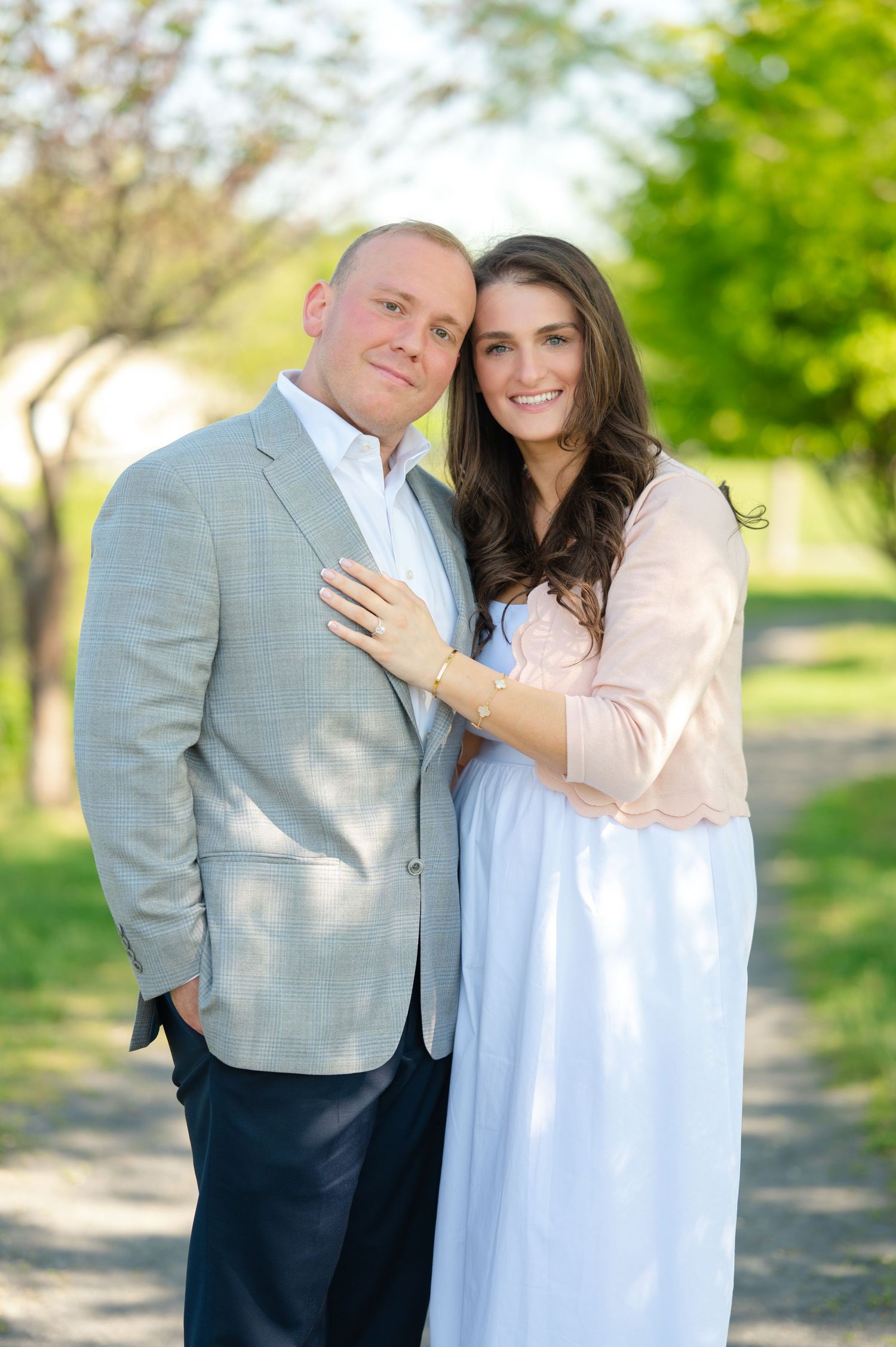 A smiling couple poses outdoors on a tree-lined path, man in gray blazer, woman in light pink and white dress.