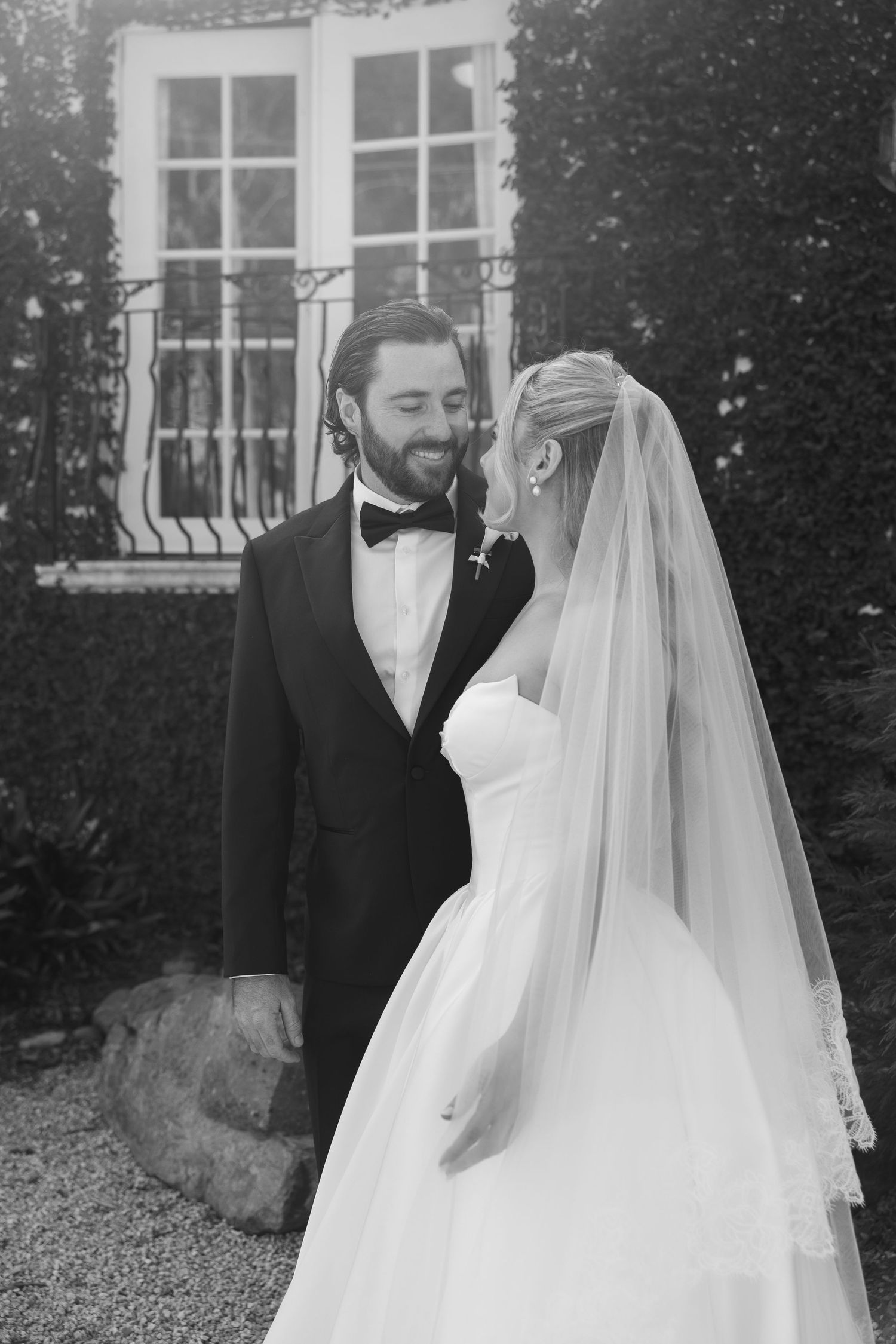 Black and white photo of a smiling bride in a strapless gown, draped in a sheer veil, seated outdoors.