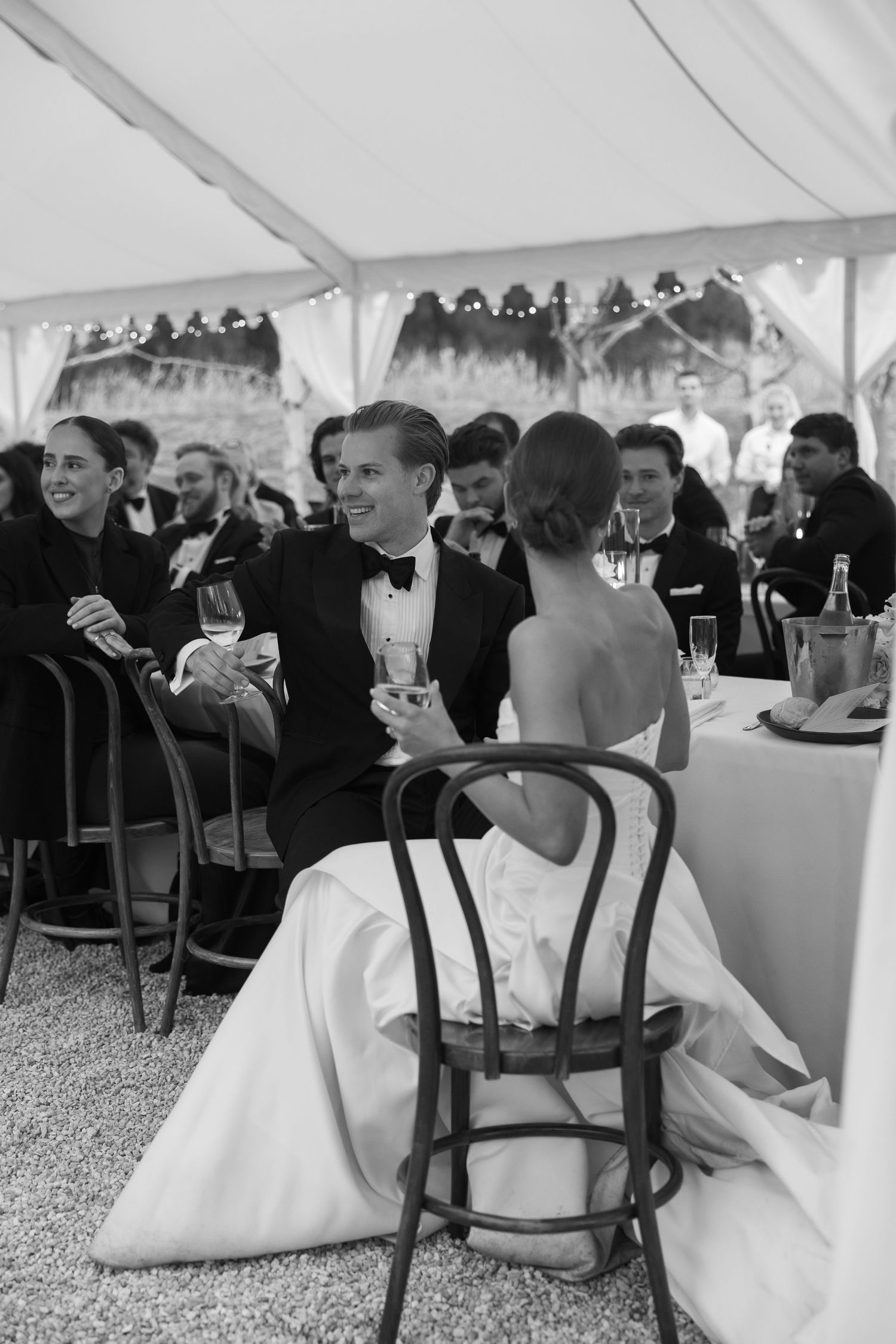 Black and white photo of a smiling bride in a strapless gown, draped in a sheer veil, seated outdoors.