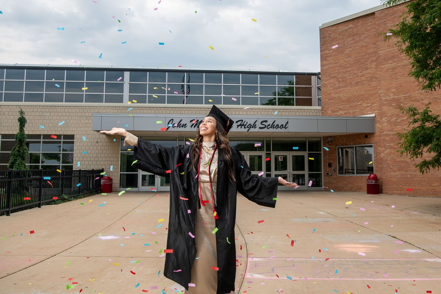 Graduate in cap and gown celebrates with colorful confetti outside John H. High School on graduation day.