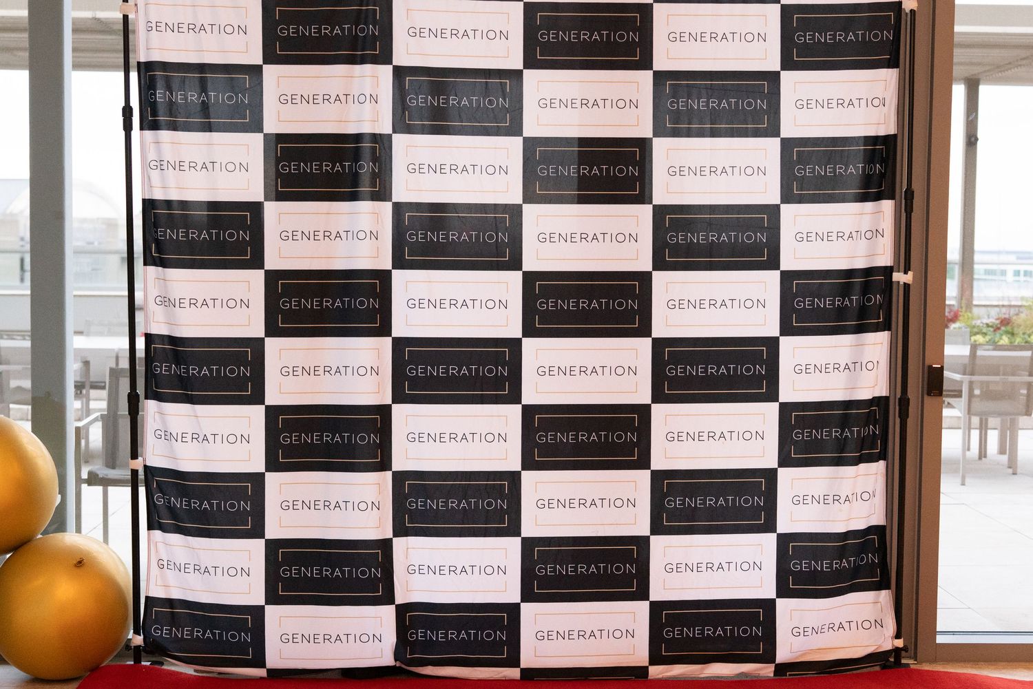 Black and white checkered Generation branded step-and-repeat backdrop with gold balloons at a corporate event.