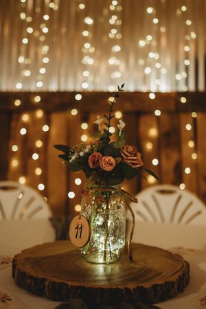 Mason jar centerpiece with flowers and fairy lights on a wood slice with bokeh background.