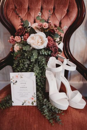 White platform bridal heels beside a floral bouquet and wedding invitation on a velvet chair.