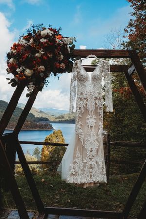 Lace wedding dress hanging on hexagonal wooden arch with mountain lake view and autumn floral arrangement.