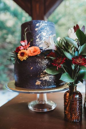 Two-tier dark navy galaxy wedding cake with gold leaf and colorful floral decorations on a glass stand.