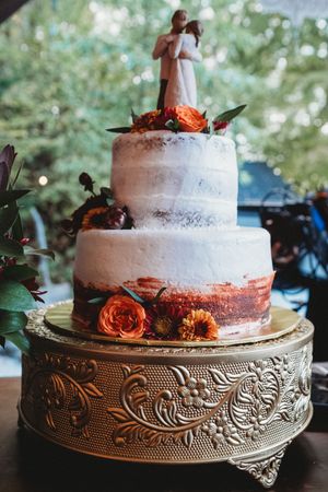 Three-tier semi-naked wedding cake with orange roses, couple topper, on ornate gold cake stand.