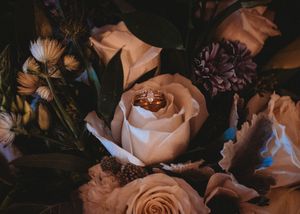 Gold wedding rings nestled among blush roses and purple flowers in a bridal bouquet.