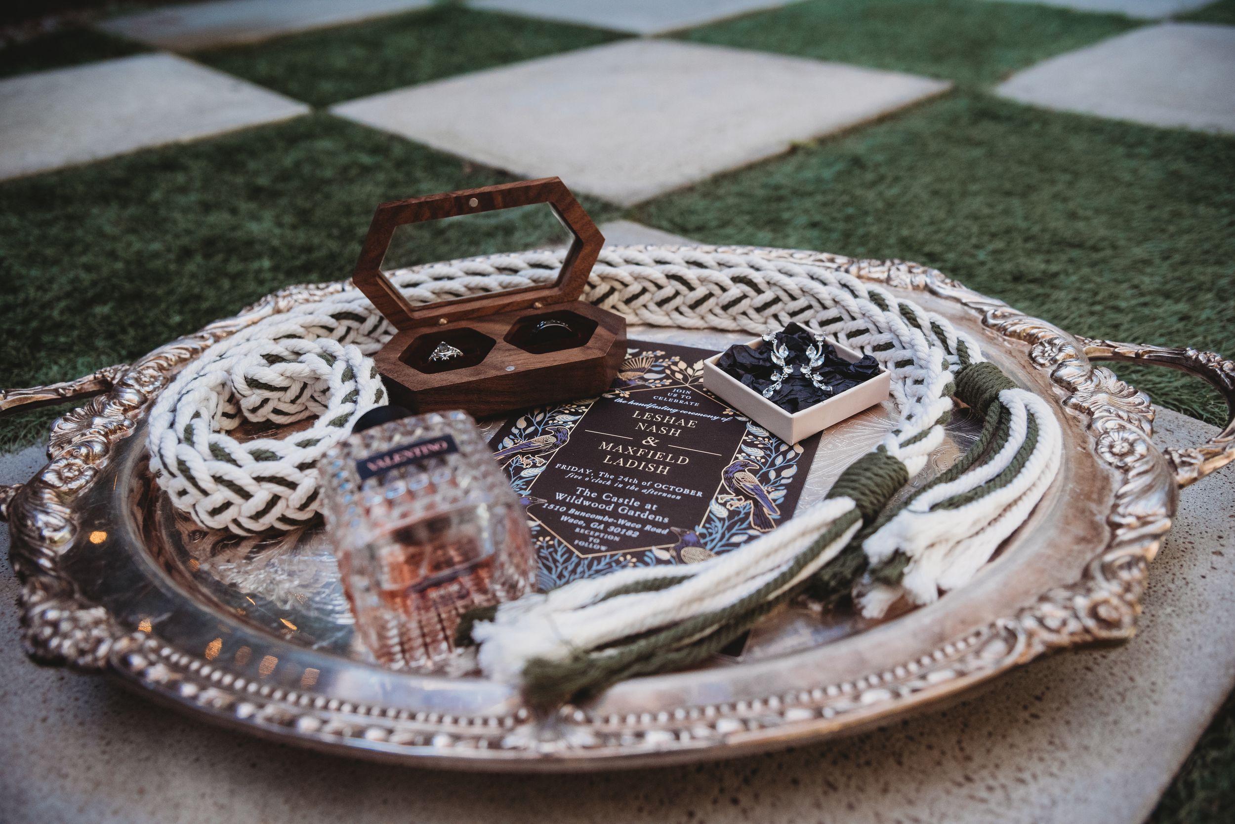 Silver tray with wedding rings, invitation, and accessories displayed on a checkered surface.