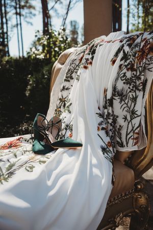 Floral embroidered white wedding dress hanging with green heels on ornate gold chair outdoors.
