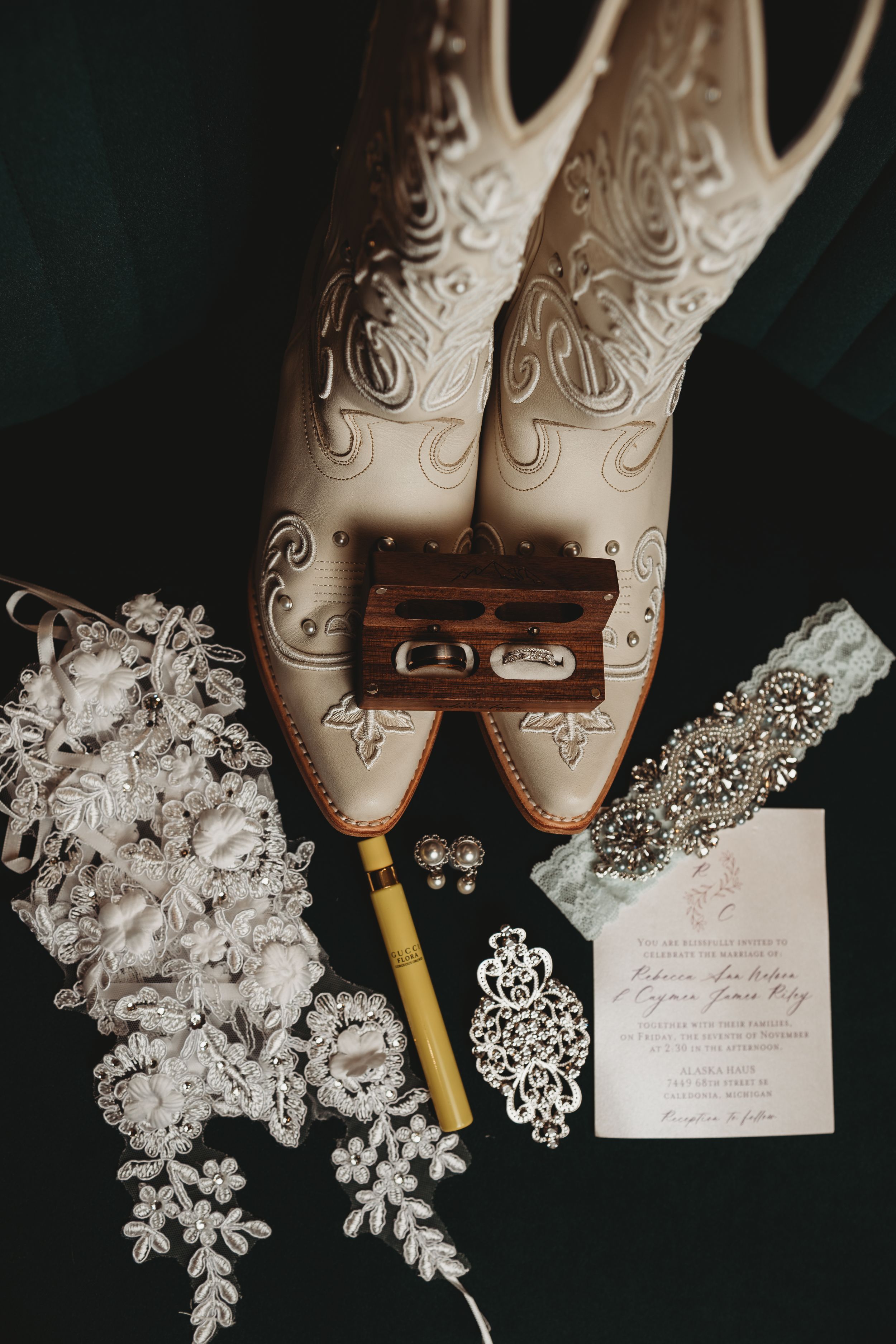Flat lay of wedding details including white cowboy boots, rings, garter, earrings, and invitation.