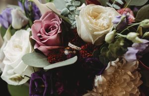 Wedding bands resting on a lush bouquet of purple, pink, and cream roses with eucalyptus.