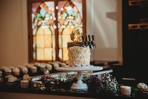 Single-tier white rosette wedding cake on a pedestal with cupcakes and stained glass backdrop.