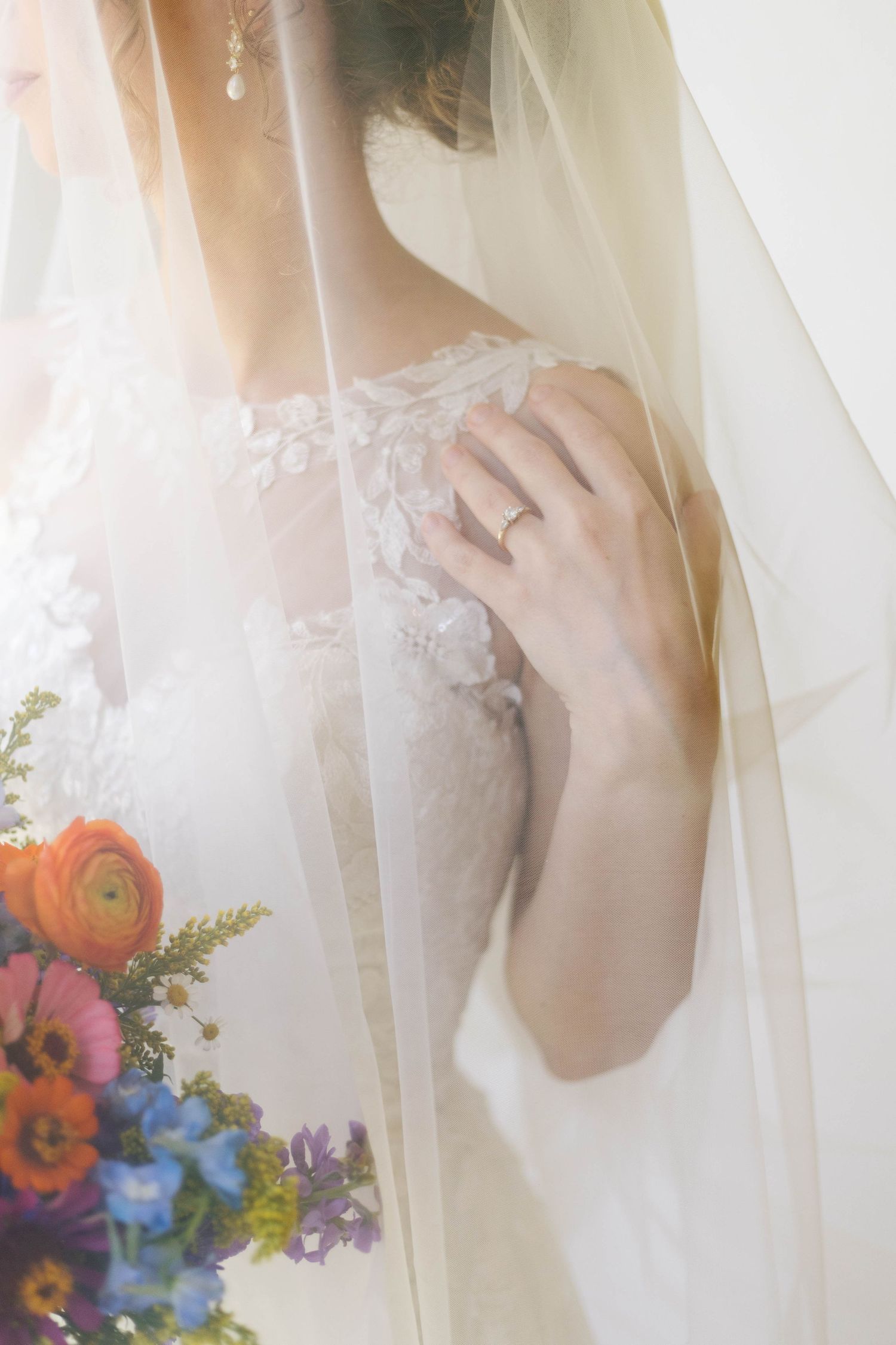 Bride in lace wedding dress with veil, wearing engagement ring, holding colorful bouquet with ranunculus and wildflowers.