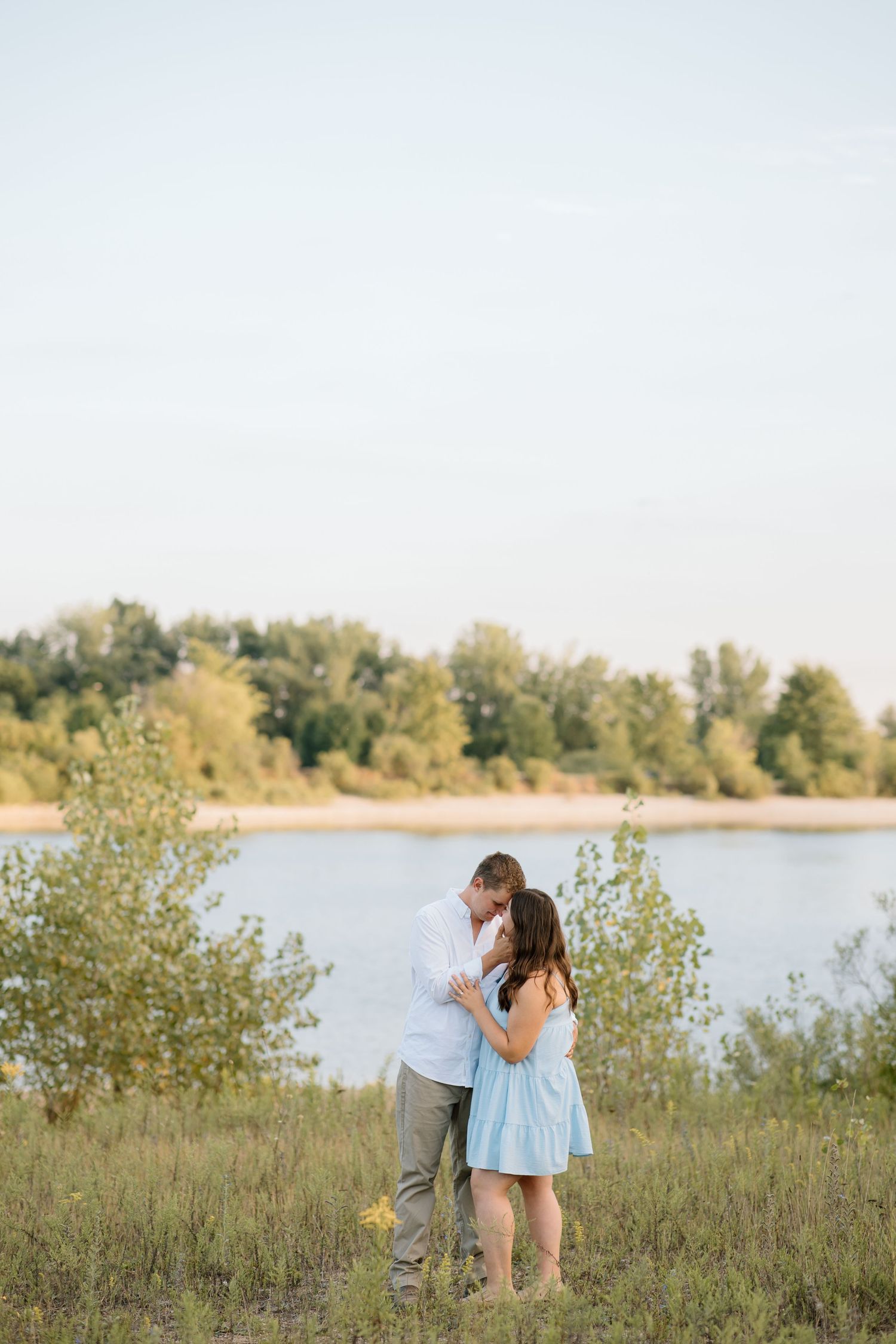 Couple sharing a romantic moment by a serene river, surrounded by lush greenery at golden hour.