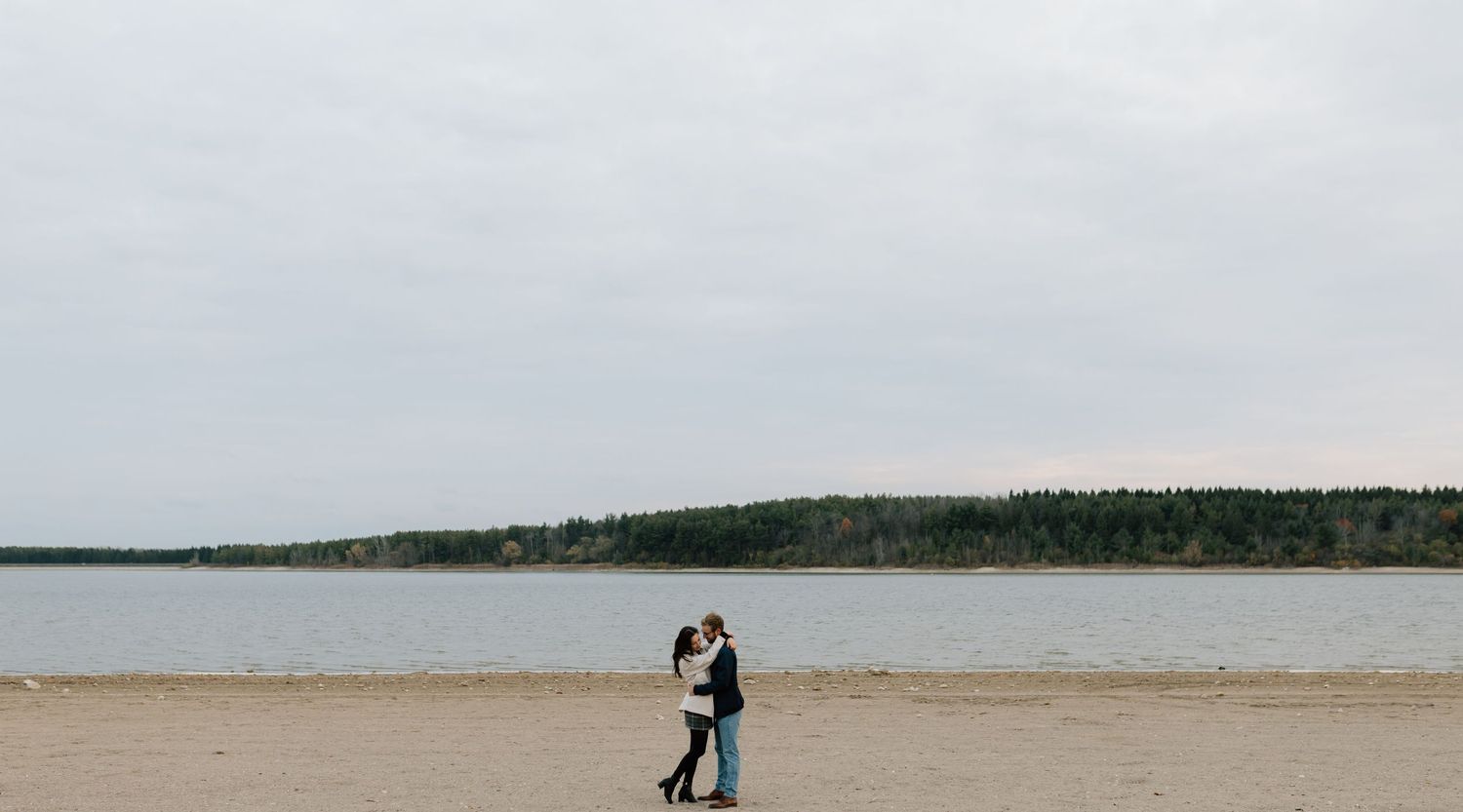 Couple embracing on a sandy beach shore with calm water and tree line under overcast skies.