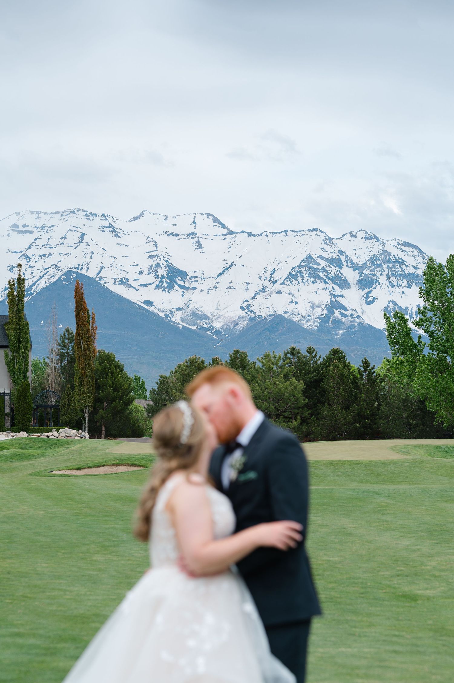 Bride and groom share a kiss on a golf course with snow-capped mountains in the background.