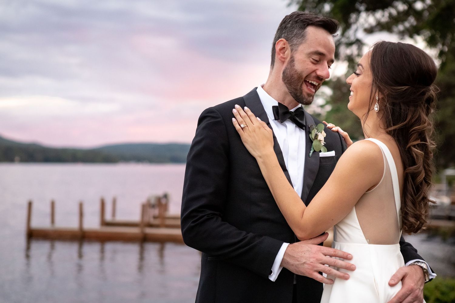 Bride and groom embrace at sunset by lakeside dock at wedding venue.