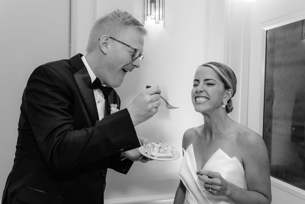Black and white photo of a groom feeding wedding cake to a laughing bride at their wedding reception.