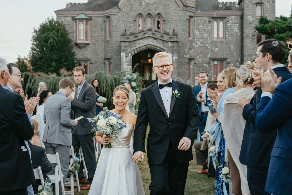 Bride and groom walk hand-in-hand down the aisle outside a stone castle venue surrounded by cheering wedding guests.