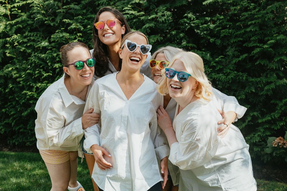Five women in white shirts and colorful heart-shaped sunglasses laughing together outdoors in a lush green garden.