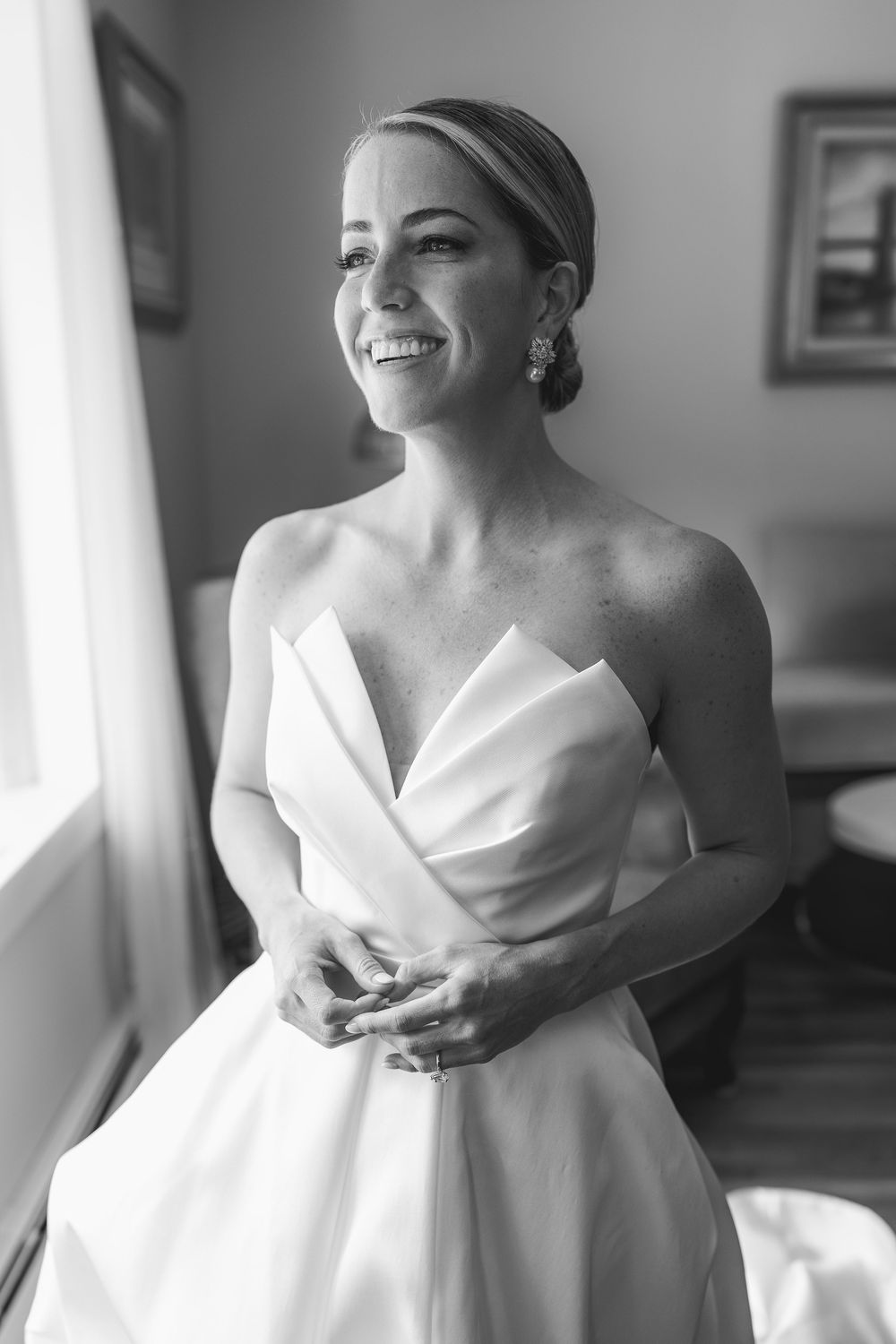 Black and white photo of a smiling bride in a strapless wedding gown standing by a window on her wedding day.