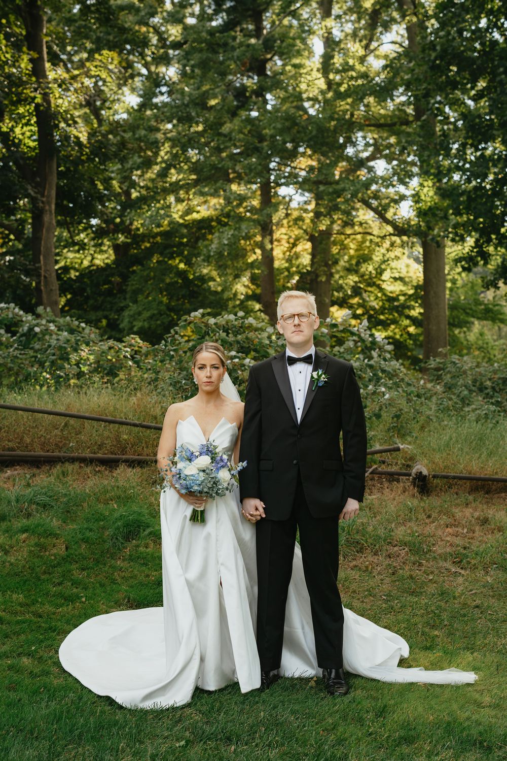 Bride in white gown holding blue bouquet stands with groom in black tuxedo outdoors among lush green trees.