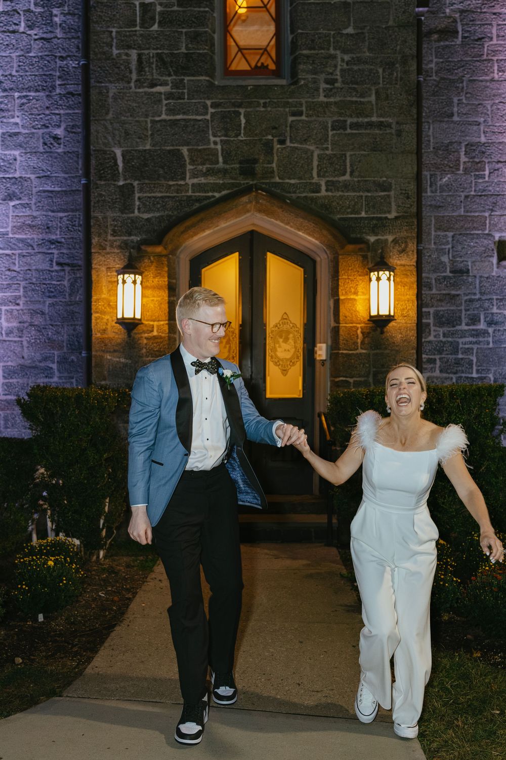 Joyful newlyweds hold hands outside a stone church at night, bride in white jumpsuit and groom in blue tuxedo jacket.