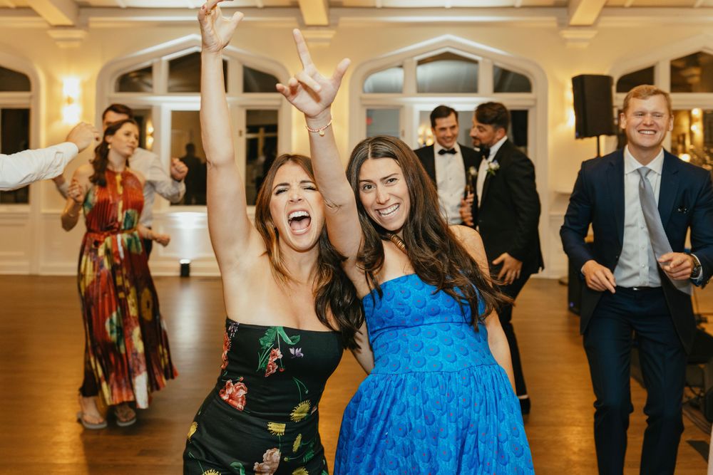 Two women dancing joyfully at a wedding reception, one in floral dress, other in blue gown, guests in formal attire behind them.