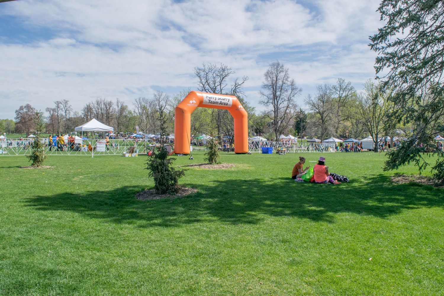 Orange inflatable arch gate at an outdoor park event with people relaxing on green grass under a sunny blue sky.