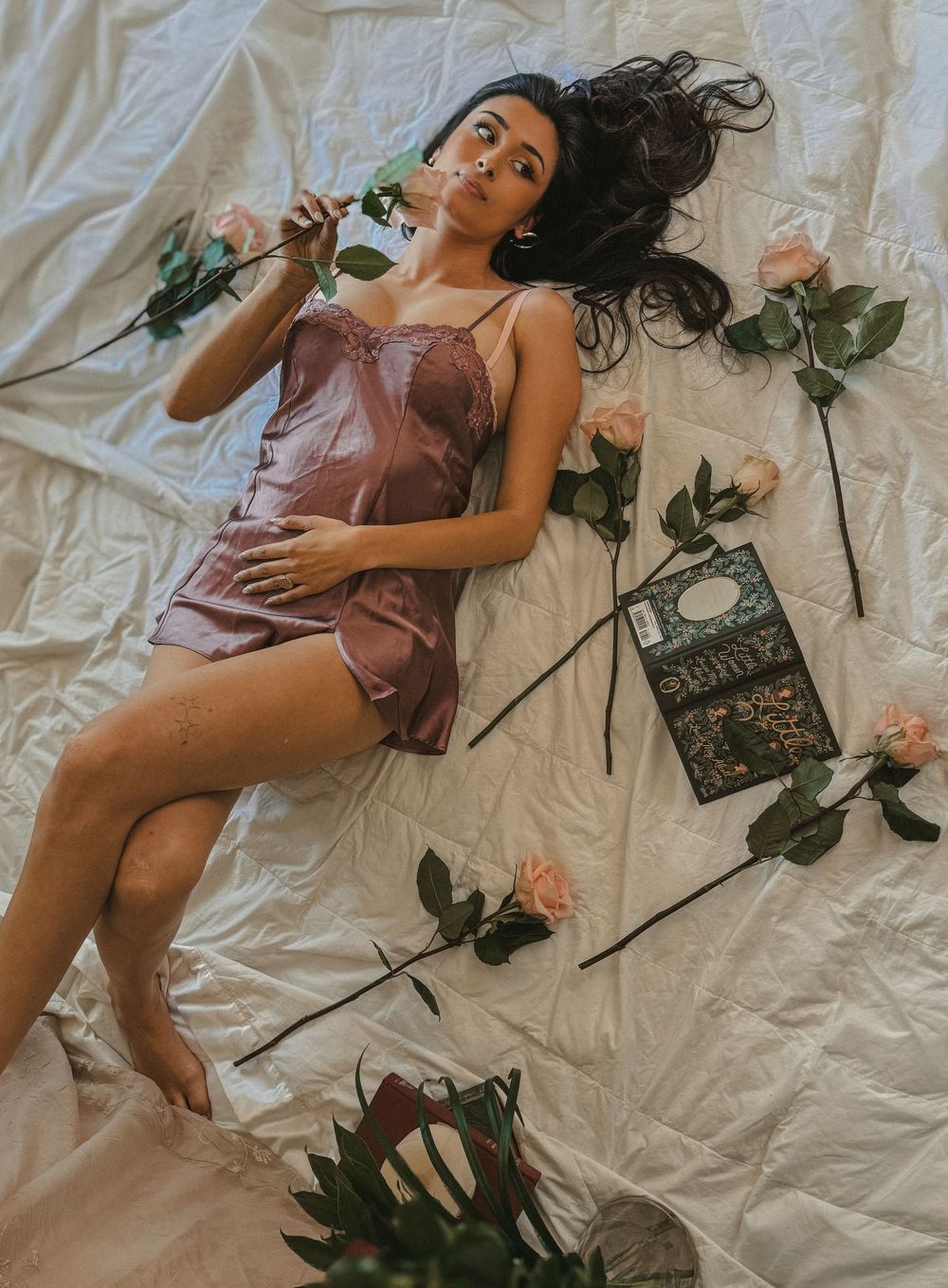 Dark-haired woman in silk slip dress lying on white sheet surrounded by long-stemmed pink roses.