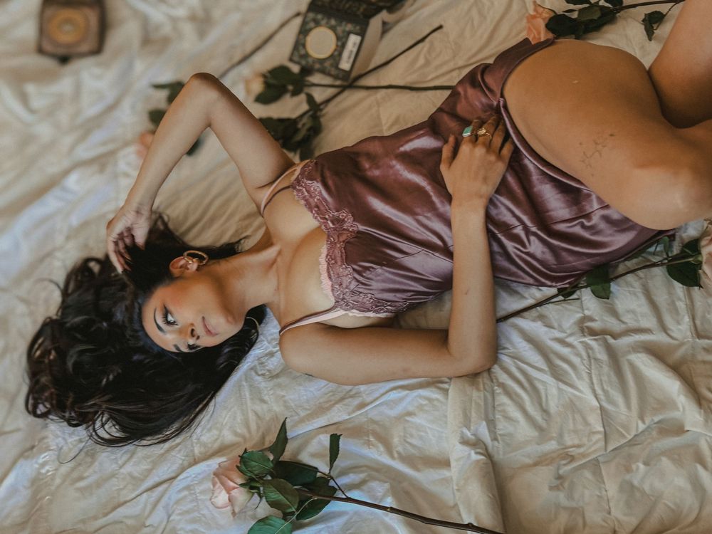 Woman in mauve silk lingerie lying on white bed with scattered roses in moody boudoir setting.