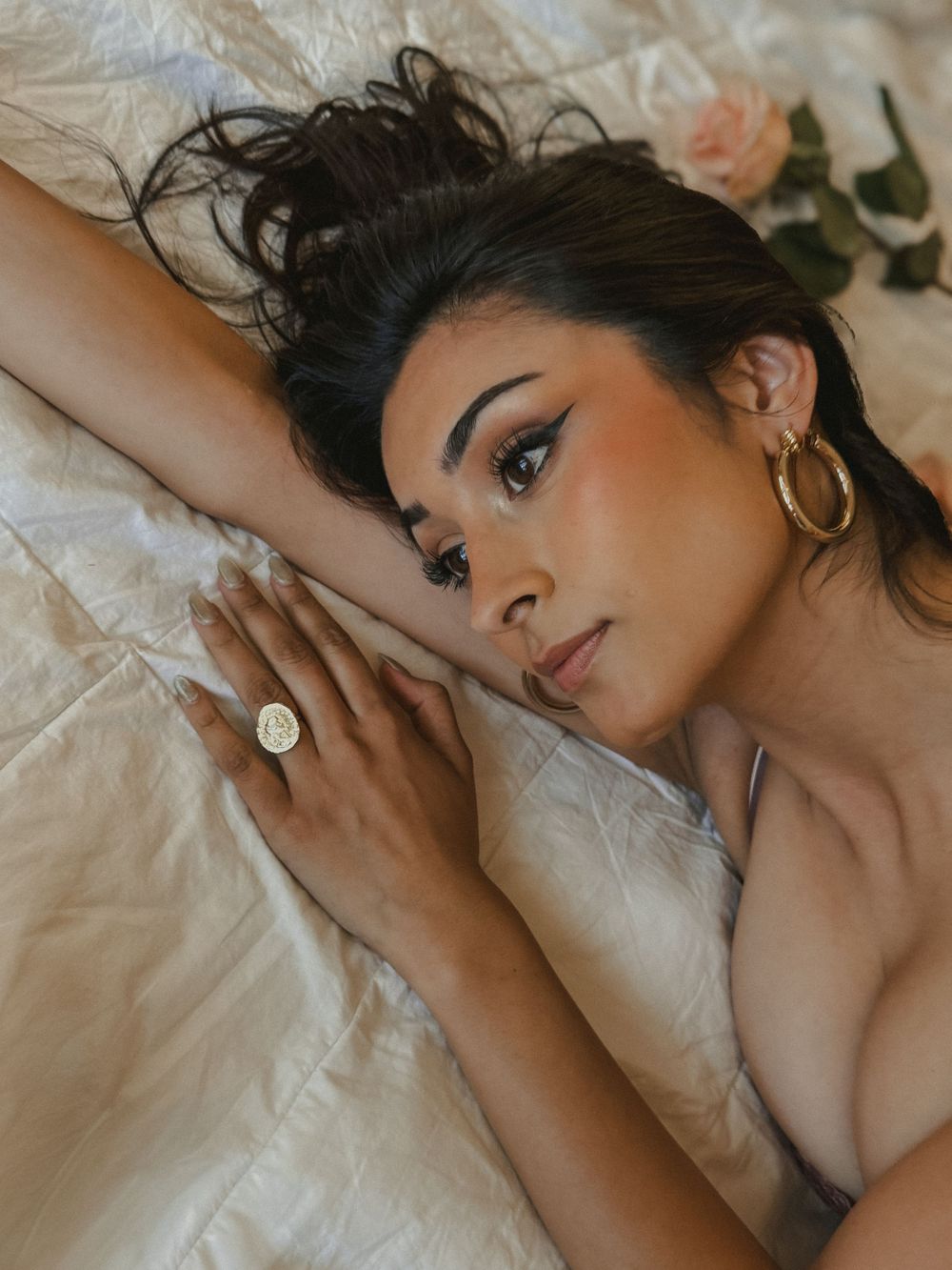 Close-up of woman with bold eye makeup lying on white bed wearing gold hoop earrings and signet ring.