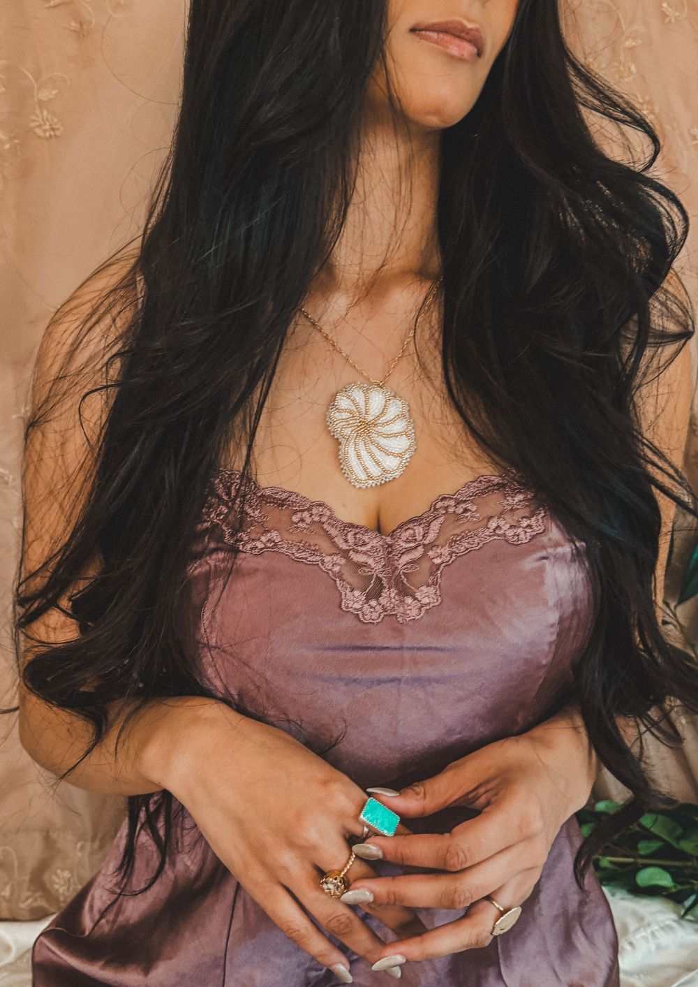 Woman with long dark hair wearing a mauve silk slip dress and large turquoise ring with nautilus shell necklace.