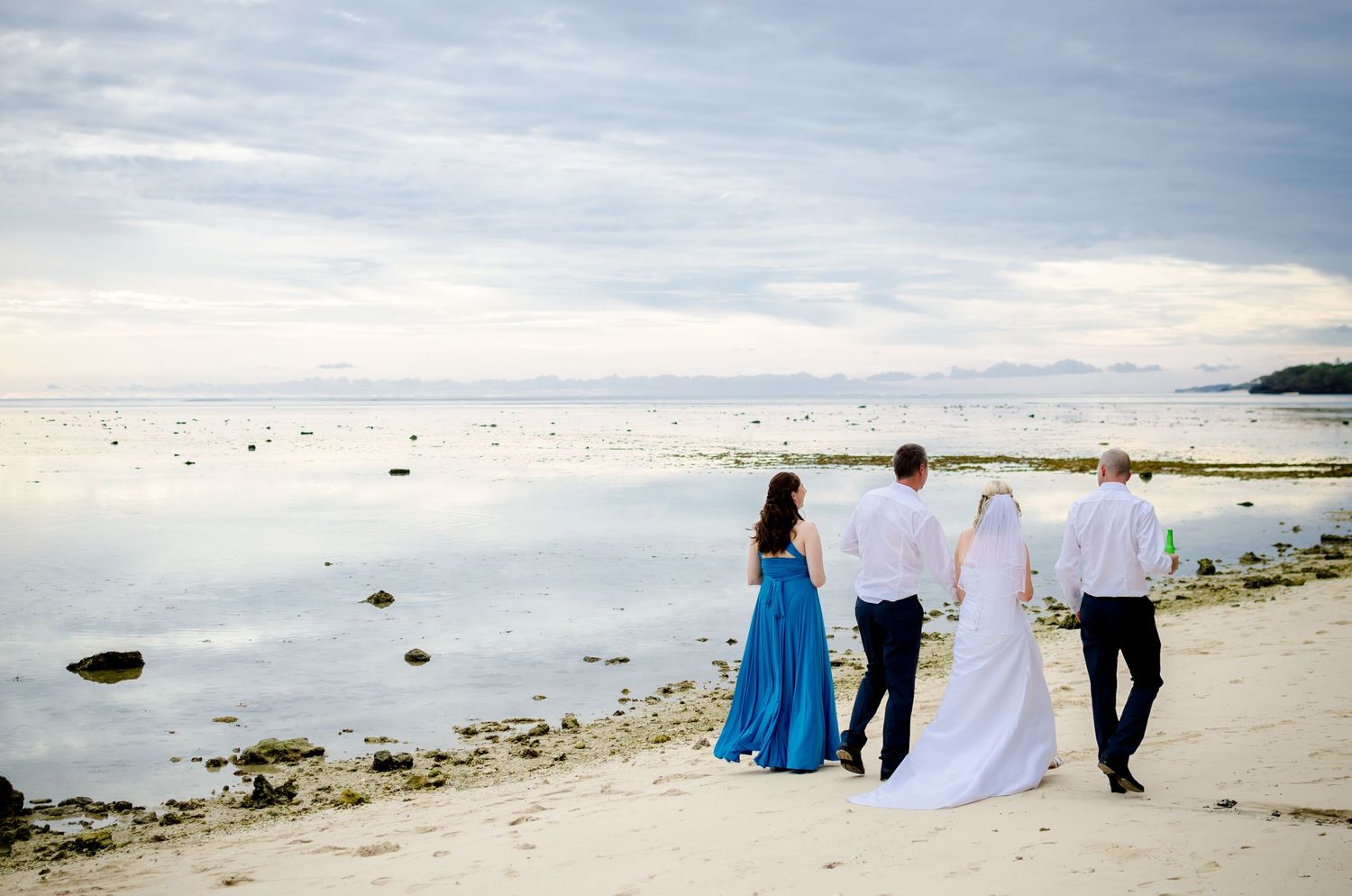 Two couples in formal attire walk along a serene beach shoreline, with calm ocean waters reflecting the cloudy sky.