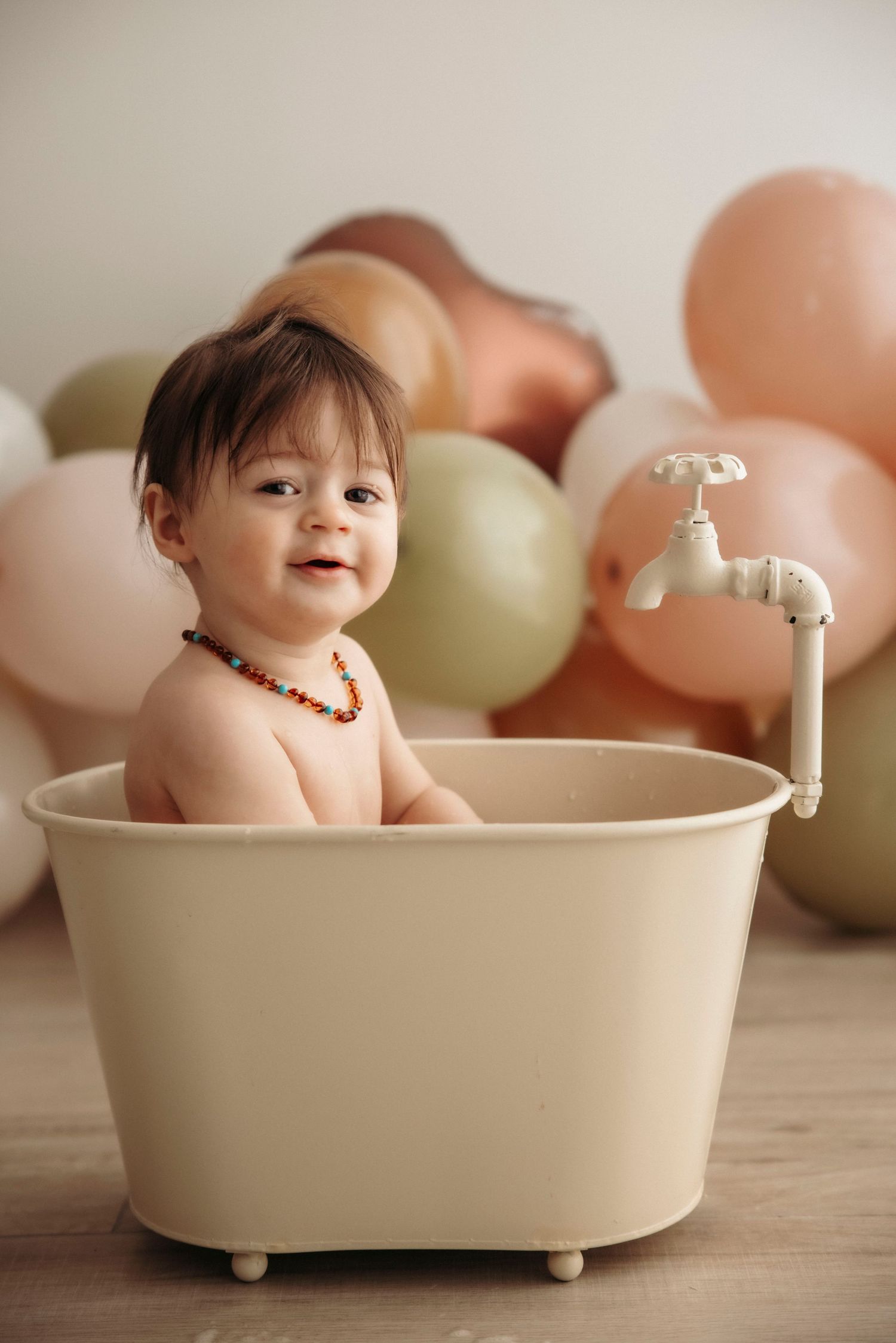 Smiling baby sitting in a small cream bathtub with colorful balloons in the background, wearing a beaded necklace.