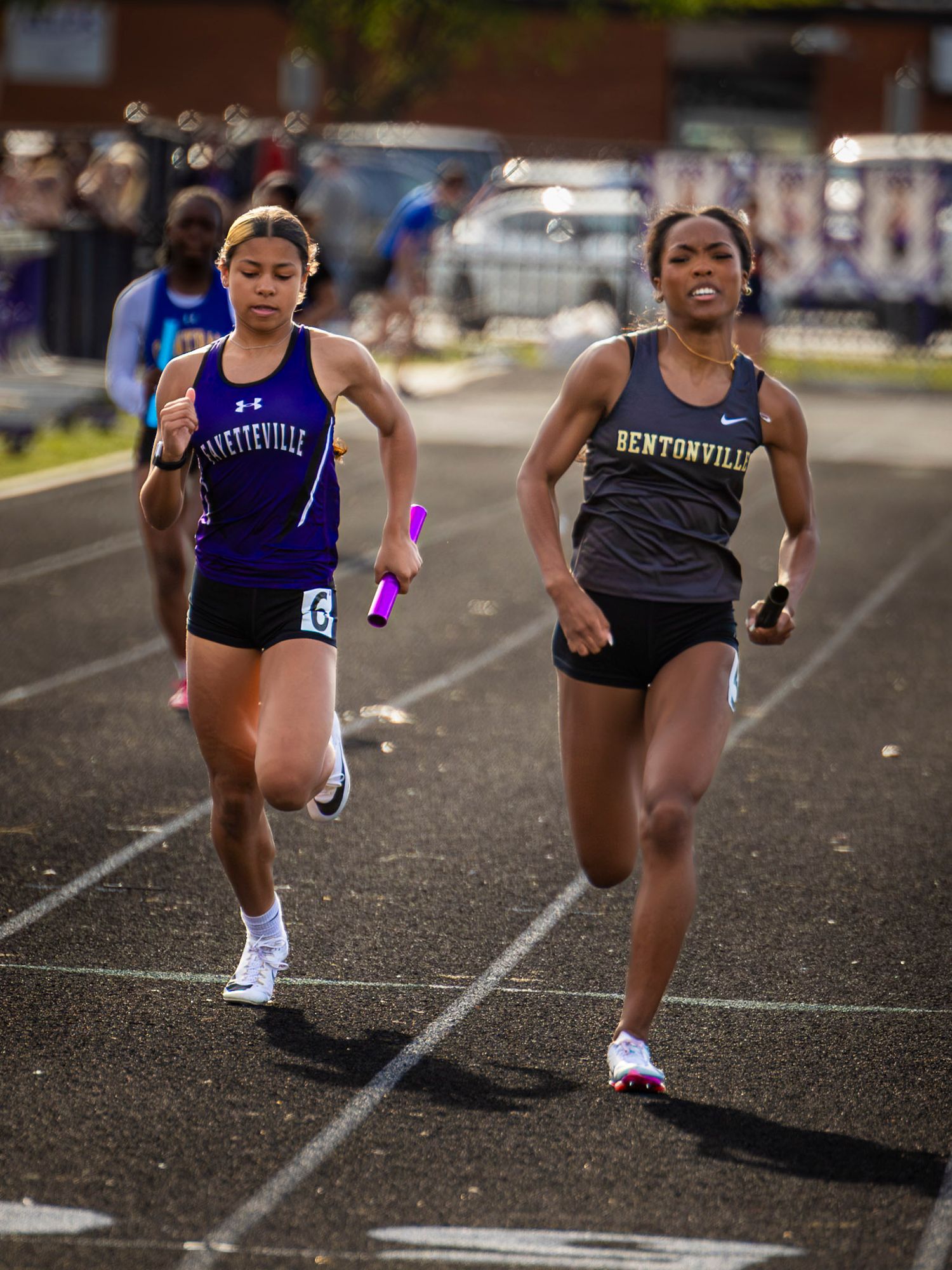 Two female athletes compete in a relay race on a track, wearing Fayetteville and Bentonville uniforms.
