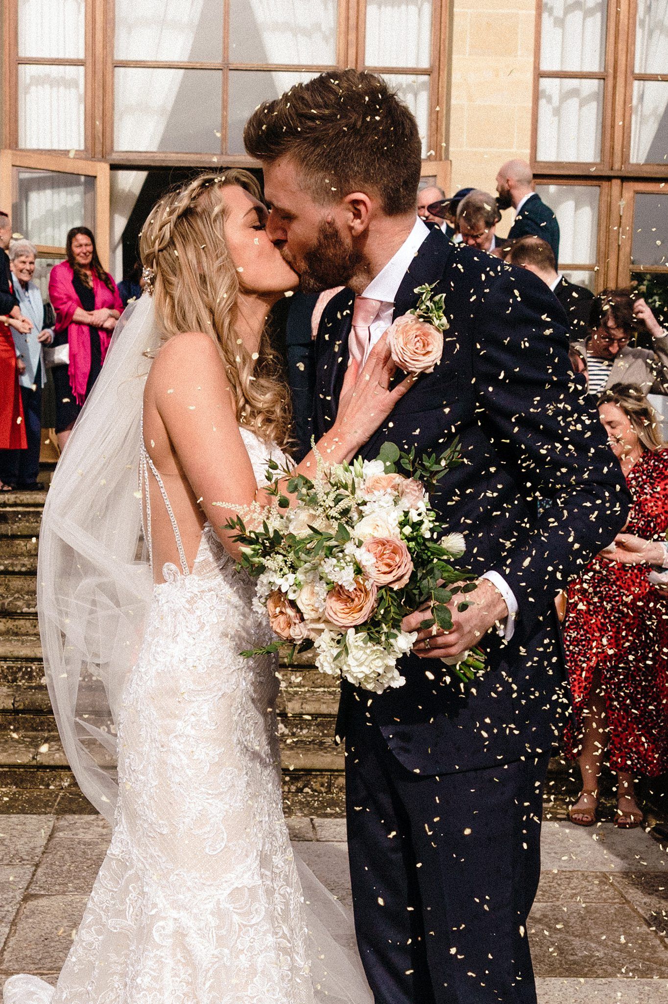 Bride and groom share a kiss surrounded by confetti, bride holding a peach bouquet outside a venue with guests watching.