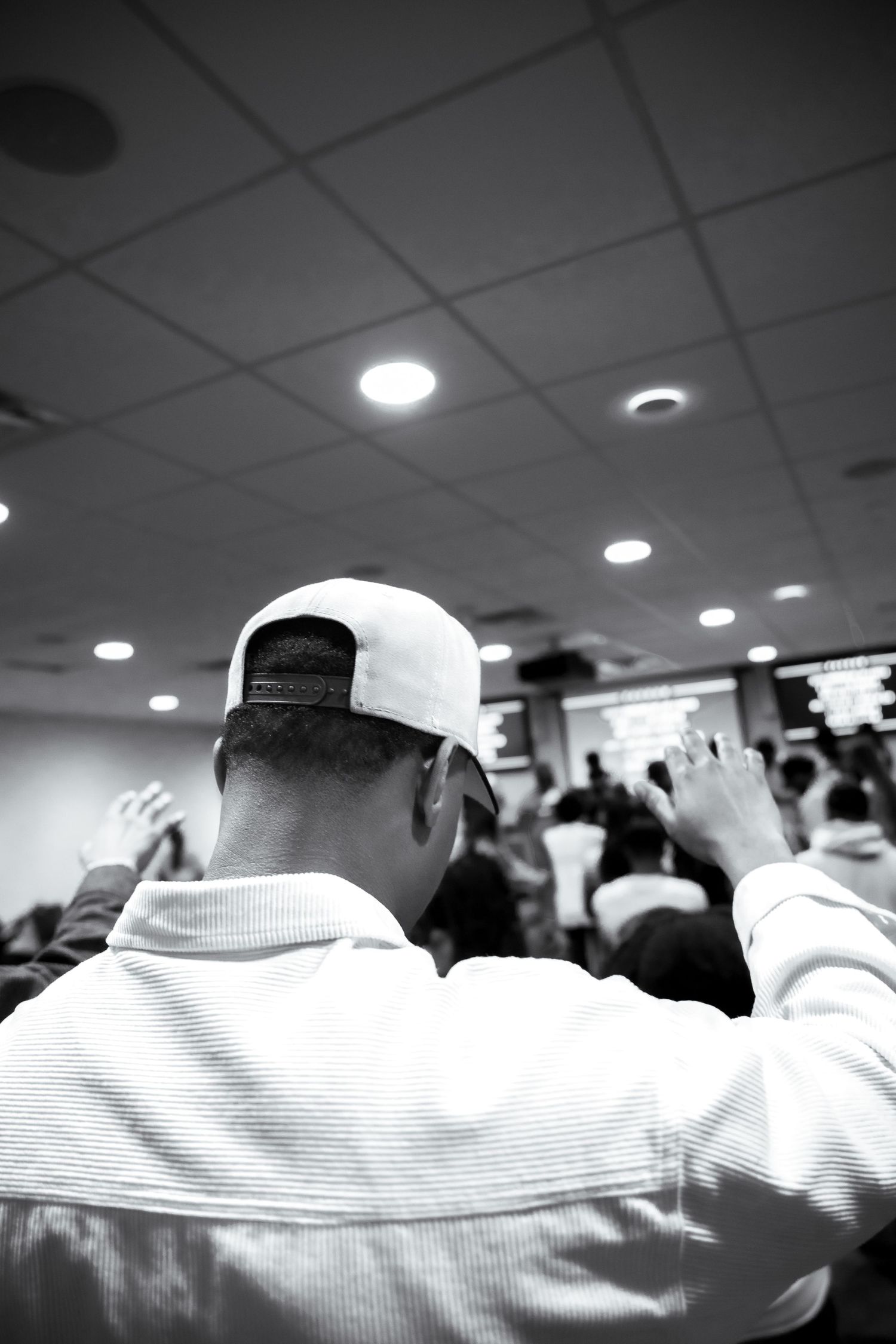 Black and white photo of a man in a white cap raising his hands in worship at a crowded indoor church service.