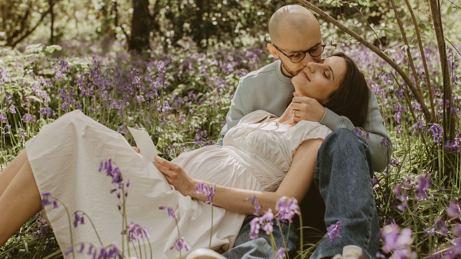 Couple sharing a tender moment in a bluebell field, pregnant woman in white dress resting on partner.
