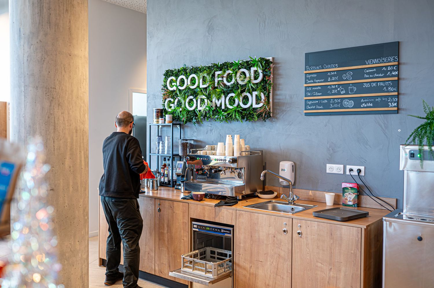 Une personne se tient à un comptoir de cuisine moderne sous un panneau néon vert "Good Food Good Mood" dans un café élégant.