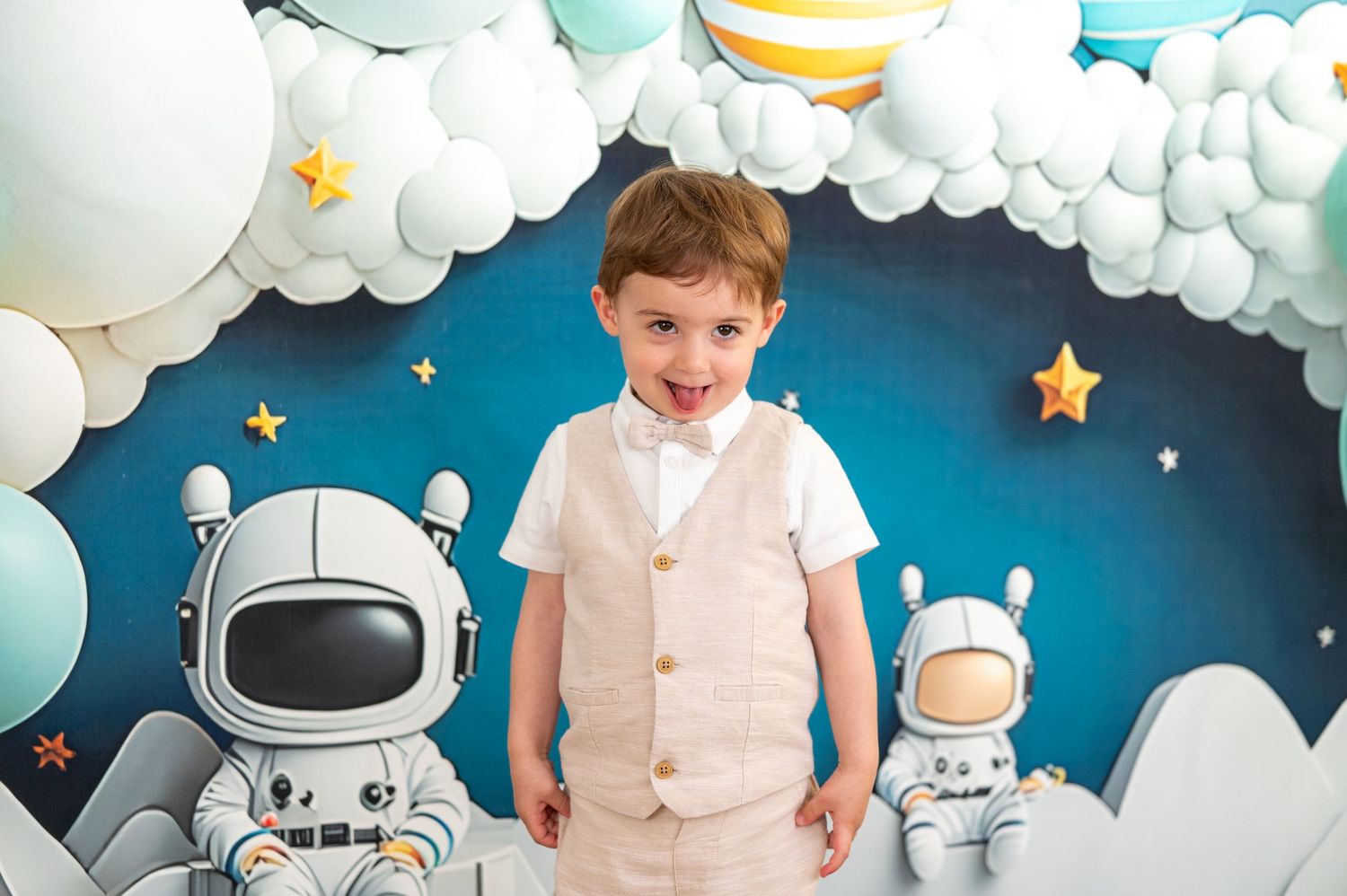 Tout-petit garçon en gilet beige souriant à une fête d'anniversaire sur le thème de l'espace avec une toile de fond d'astronaute et des nuag