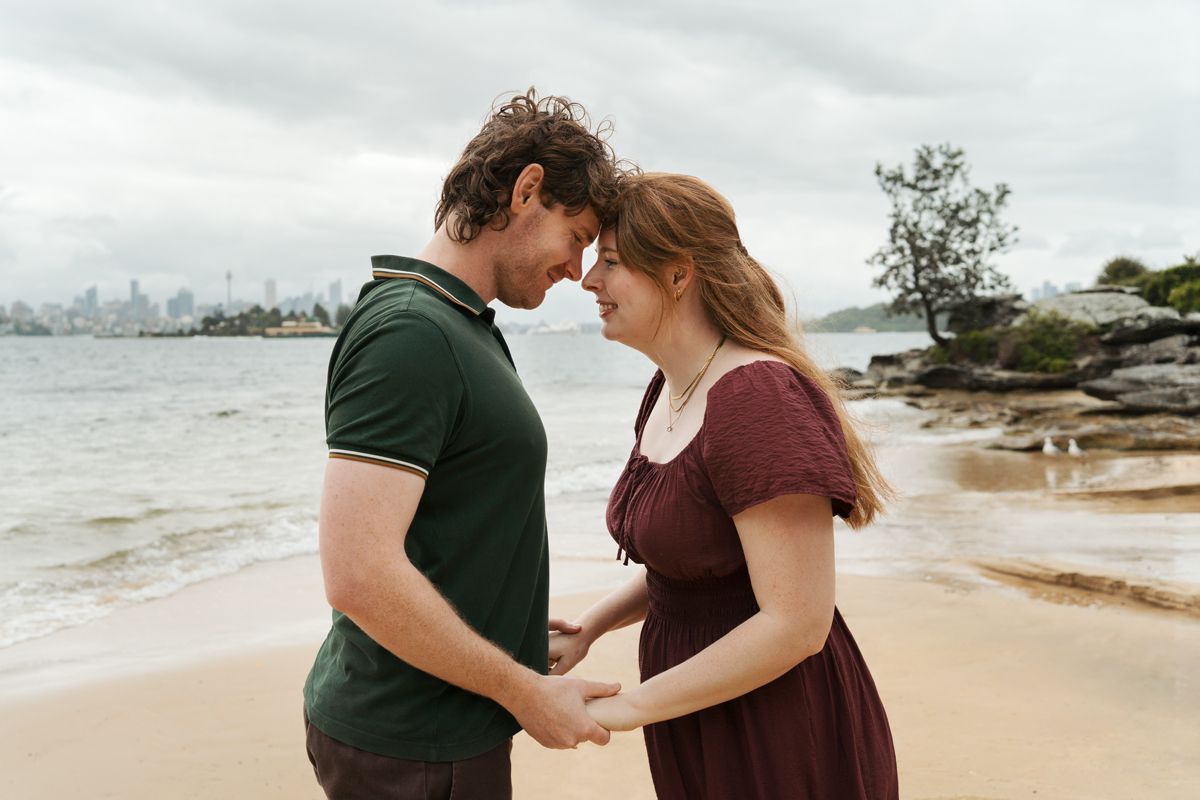 Couple holding hands on a sandy beach, sharing an intimate moment with a city skyline in the background during proposal photoshoot