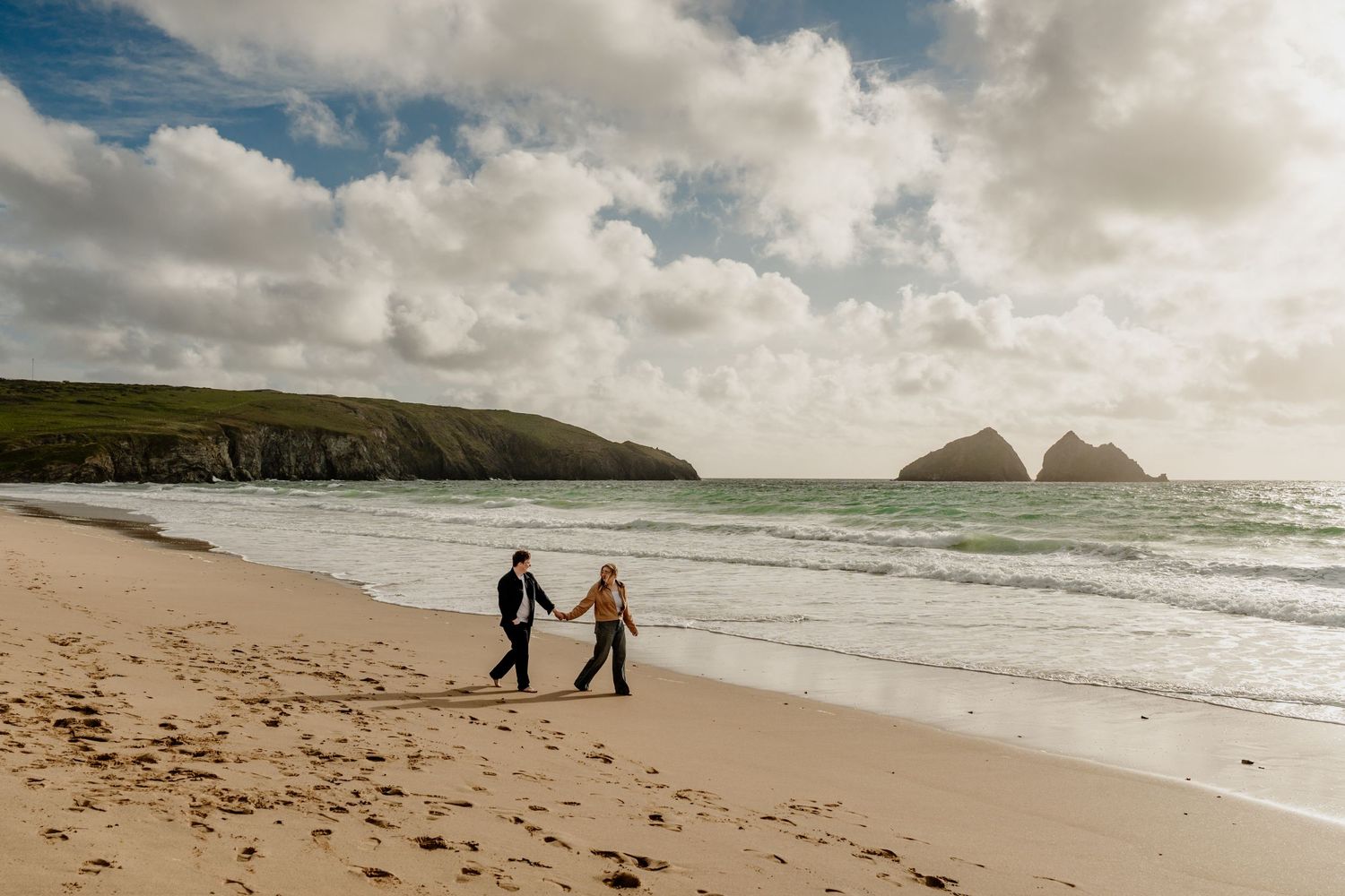 Couple walking hand in hand on the beach at Holywell Bay with dramatic cloudy skies in the background.