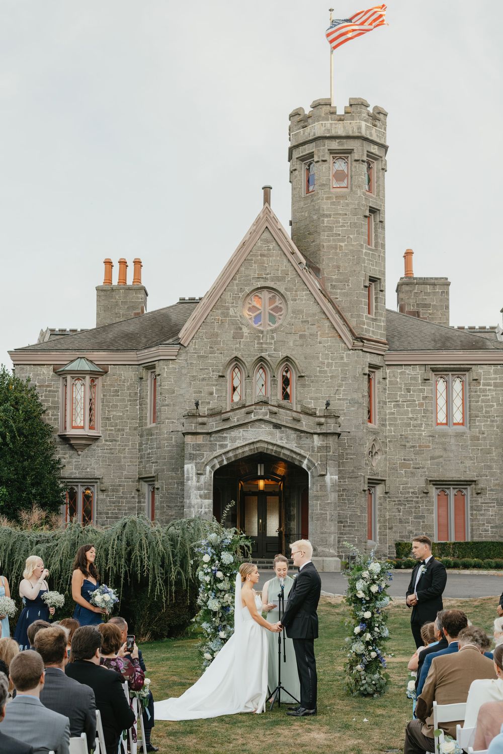 A wedding ceremony takes place in front of a grand stone castle with Gothic architecture and multiple turrets.