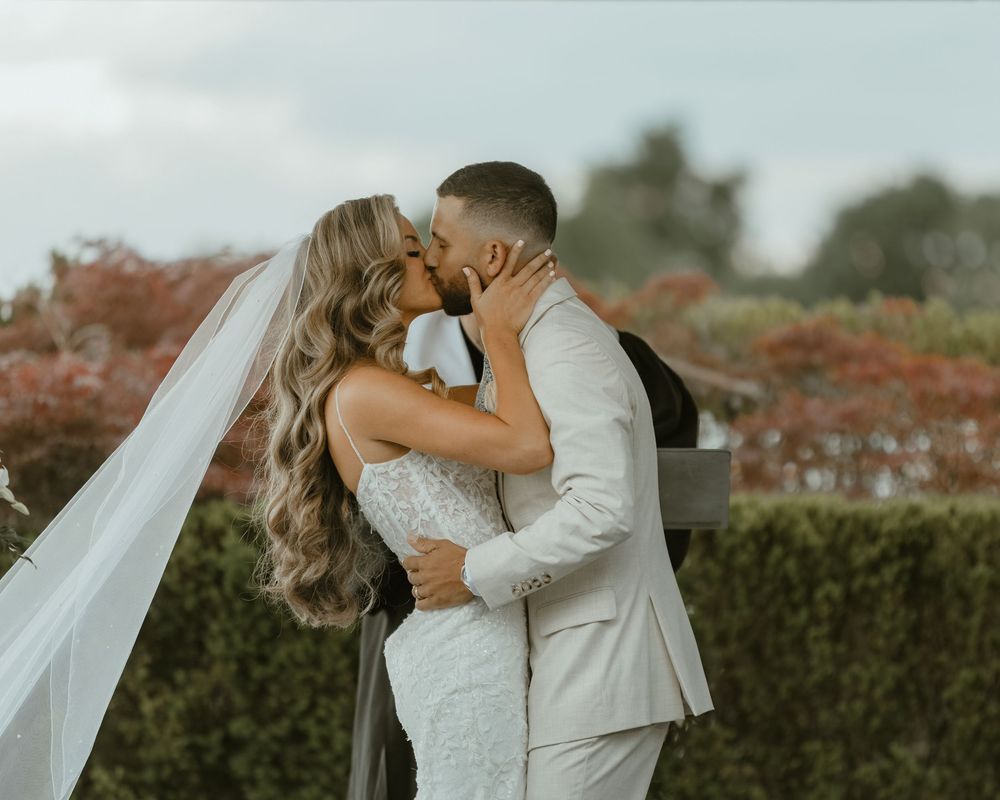 A couple shares a romantic kiss on their wedding day with red autumn foliage and hedges in the background.