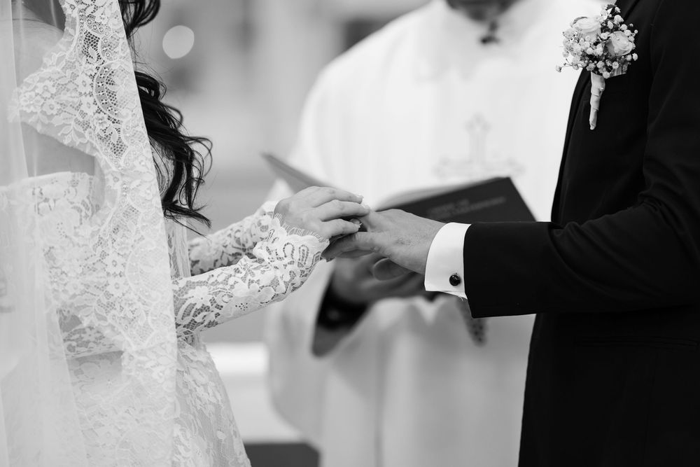 Black and white closeup of wedding ceremony showing exchange of rings with bride in lace dress and groom in dark suit.